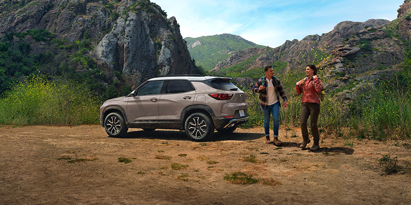 A beige 2026 Chevrolet Trax parked in a mountainous landscape, with two people walking nearby, enjoying an outdoor adventure.