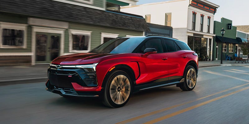 Red Chevrolet Blazer EV with a black roof driving through a downtown street lined with storefronts.
