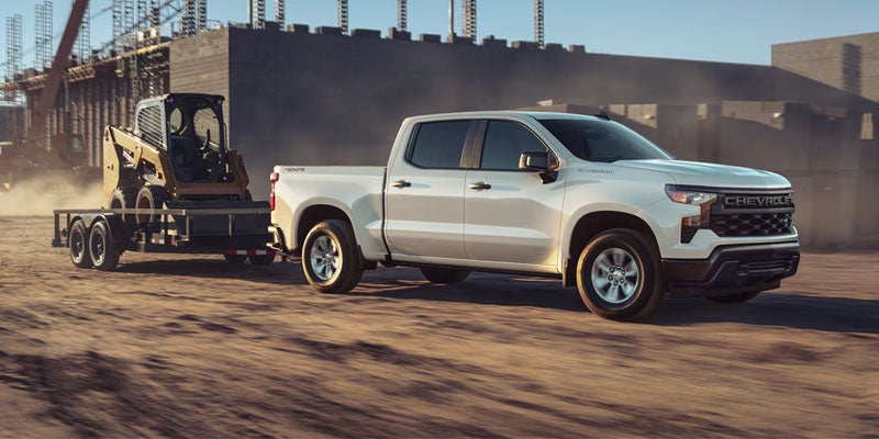 White Chevrolet Silverado 1500 crew cab pickup truck towing a flatbed trailer with a skid steer loader at a construction site.
